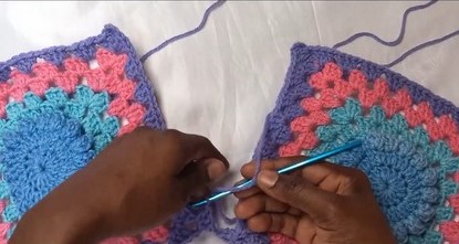 Hands joining two pink, teal, and purple granny squares side by side with a teal hook on a white surface.