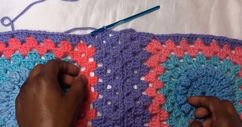 Hands working a purple joining row along the seam between two rows of multicolor granny squares with a teal hook.
