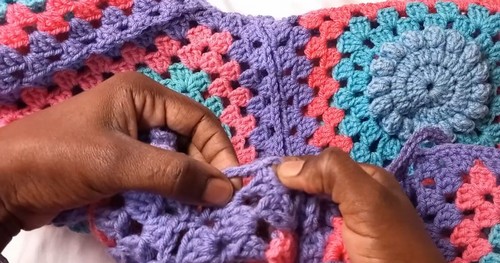 Hands guiding yarn through the corner join of a row of pink, teal, and purple granny squares on a white surface.