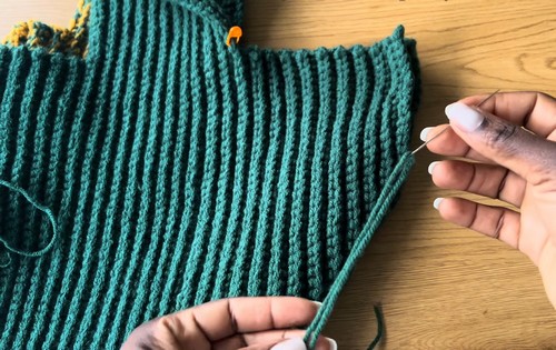 Hands seaming the side of a teal ribbed crochet sweater body showing the vertical rib texture and stitch markers