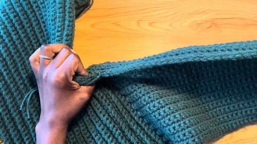 Hand seaming the shoulder edge of a teal ribbed crochet sweater panel on a wooden surface
