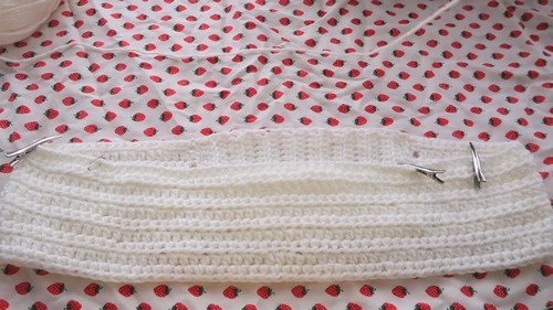 White ribbed crochet top band pinned flat on a strawberry print fabric surface with two metal clips holding the edges.