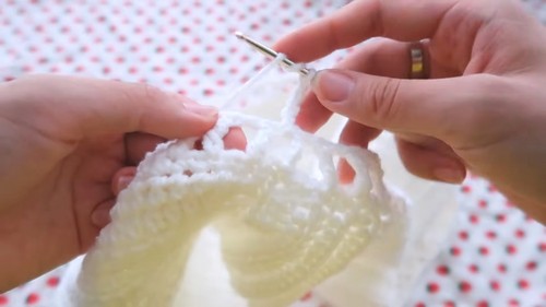 Hands working a silver hook through the lace edge of a white crochet top piece on a red dotted fabric surface.