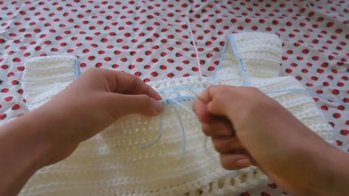 Hands threading a thin blue ribbon through the center front neckline of a flat white crochet square neck top.