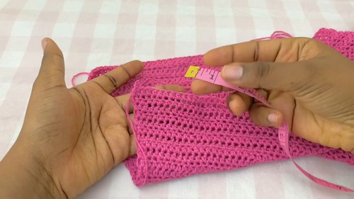 Pink crochet swatch being measured with a yellow tape measure on a gingham surface