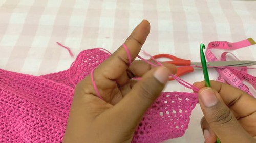 Pink crochet fabric with a green hook and scissors beside a measuring tape on a gingham background