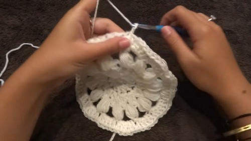 Hands building a circular mandala motif in white yarn with puff stitch petals using a blue hook