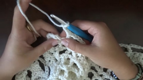 Hands working a blue hook into the border rounds of a cream lace motif with open shell and chain space detail
