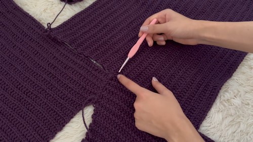Purple ribbed sweater panel laid flat on a white surface with a pink crochet hook joining a sleeve