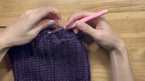 Purple ribbed crochet fabric in progress on a wooden surface with a pink hook