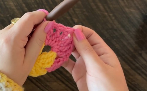 Hands crocheting the center rounds of a pink and yellow granny square with a dark wood hook.
