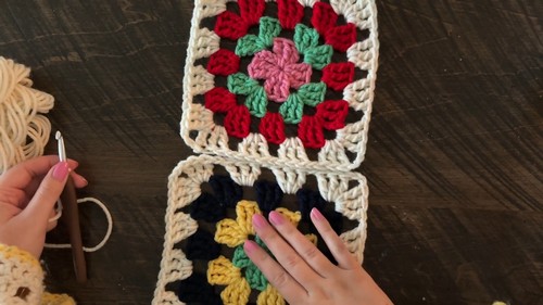 Two completed granny squares in multicolor and cream laid flat with hands and a hook positioned to join them.