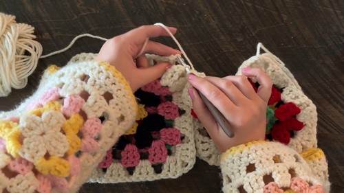 Hands joining two cream bordered granny squares with a tapestry needle on a dark wood surface.