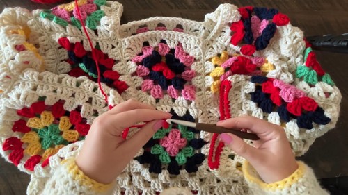 Hands using a dark hook to join a row of multicolor granny squares along a red joining seam on a dark surface.