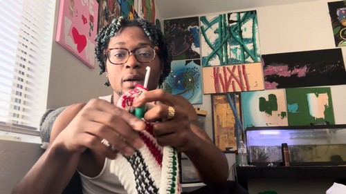 Working a red and white striped panel of the vest with a green crochet hook while seated indoors