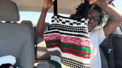 Holding up the finished black, white, red and green striped vest inside a car to show the full length and shoulder straps