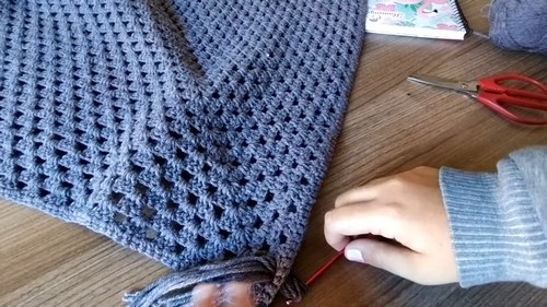 Attaching grey fringe to the corner of the finished poncho with a red hook, scissors and a notebook on the table